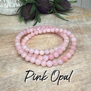 May include: Pink opal beaded bracelet. The bracelet is made of round, light pink beads. The text "Pink Opal" is written in a cursive font below the bracelet. The bracelet is displayed on a wooden surface.