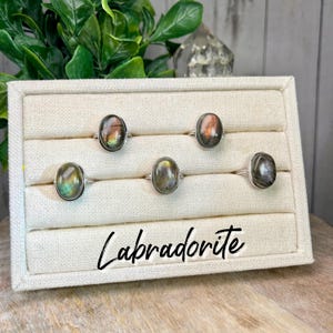 May include: Five silver rings with oval-shaped labradorite gemstones displayed on a white fabric ring display. The rings are arranged in a row with the text "Labradorite" printed below them.