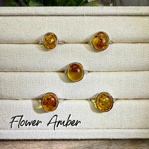 May include: Five silver rings with amber-colored stones are displayed. The amber stones have inclusions, and the rings are set against a neutral background. The text "Flower Amber" is written in cursive at the bottom.