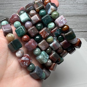 May include: A stack of bracelets made with green, brown, and red agate gemstone beads. The beads are square and round in shape.