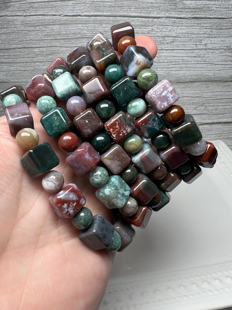 May include: A stack of bracelets made with green, brown, and red agate gemstone beads. The beads are square and round in shape.