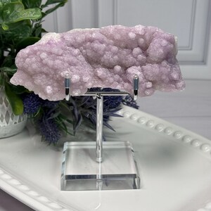May include: A light purple amethyst geode specimen displayed on a clear acrylic stand with a silver metal support. The geode has a bubbly, crystalline texture and is set against a white background with greenery.