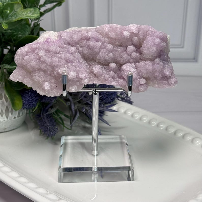 May include: A light purple amethyst geode specimen displayed on a clear acrylic stand with a silver metal support. The geode has a bubbly, crystalline texture and is set against a white background with greenery.
