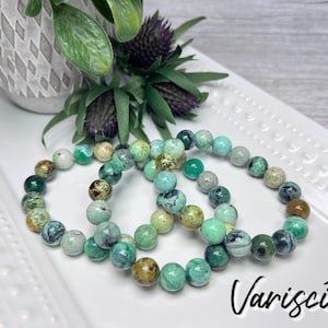 May include: Three green and blue gemstone bracelets made of variscite beads. The bracelets are stacked on a white plate.