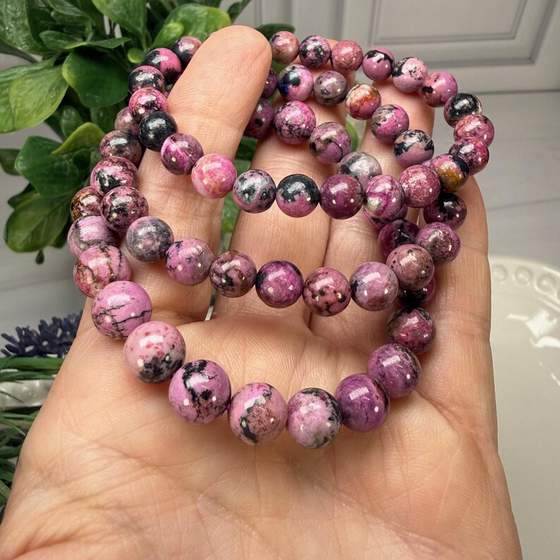 May include: Three beaded bracelets featuring pink and black gemstone beads. The bracelets are held in a hand, showcasing the varying shades of pink and the speckled black patterns within the stones. The beads appear to be approximately 0.4 inches in diameter.