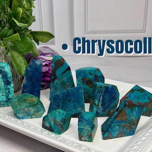 May include: A collection of raw chrysocolla mineral specimens featuring vibrant blue and green hues with dark brown matrix inclusions, arranged on a white rectangular tray. The text "Chrysocolla" is displayed in blue letters above the stones.