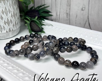 May include: A set of four bracelets made with black and gray volcano agate beads. The bracelets are stacked on a white surface. Volcano Agate.