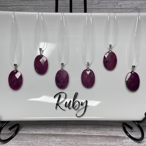 May include: Six ruby gemstone pendants on clear elastic cords, displayed on a white plate with the word "Ruby" written in black cursive.