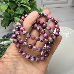 May include: Three beaded bracelets with pink and black stones. The bracelets are made of round, polished beads in varying shades of pink and black. The bracelets are held in a hand, with a blurred background of greenery and a white dish.