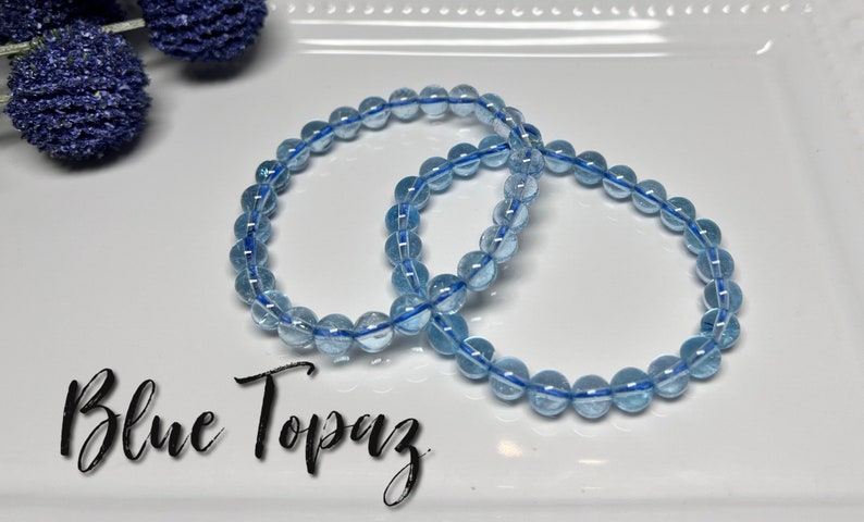 May include: Two bracelets made of blue topaz beads are shown on a white plate. The beads are strung on a blue elastic cord. The text "Blue Topaz" is written in a cursive font at the bottom of the image. Decorative blue floral accents are in the background.