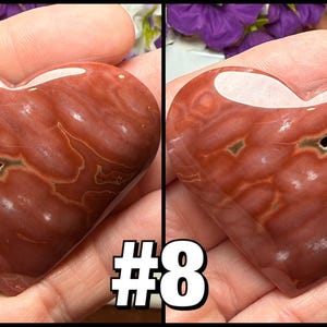 May include: A polished, heart-shaped stone with a reddish-brown hue and white veining. The stone has a natural cavity with green and white accents. The number "#8" is displayed in a white font with a black outline.
