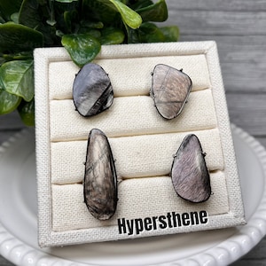 May include: Four rings with a black metal setting and a gray and black gemstone. The rings are displayed on a white fabric ring display. The text "Hypersthene" is printed on the display.