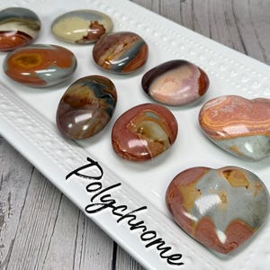 May include: A collection of polished polychrome jasper stones in various shapes, including ovals and hearts, displayed on a white rectangular tray. The stones feature a mix of earthy tones like red, brown, gray, and cream.