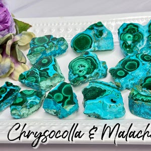 May include: A collection of raw chrysocolla and malachite mineral specimens featuring vibrant turquoise and deep green circular patterns, arranged on a white rectangular tray with a beaded edge, accompanied by a purple flower.
