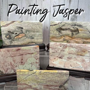 May include: Five rectangular Painting Jasper stones on wooden stands. The stones display a variety of colors, including beige, pink, and gray, with intricate patterns. The text "Painting Jasper" is written in a cursive font.