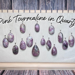 May include: Pink Tourmaline in Quartz pendants on silver chains displayed on a wooden board. The pendants are various shapes and sizes, with a mix of pink and white colors.