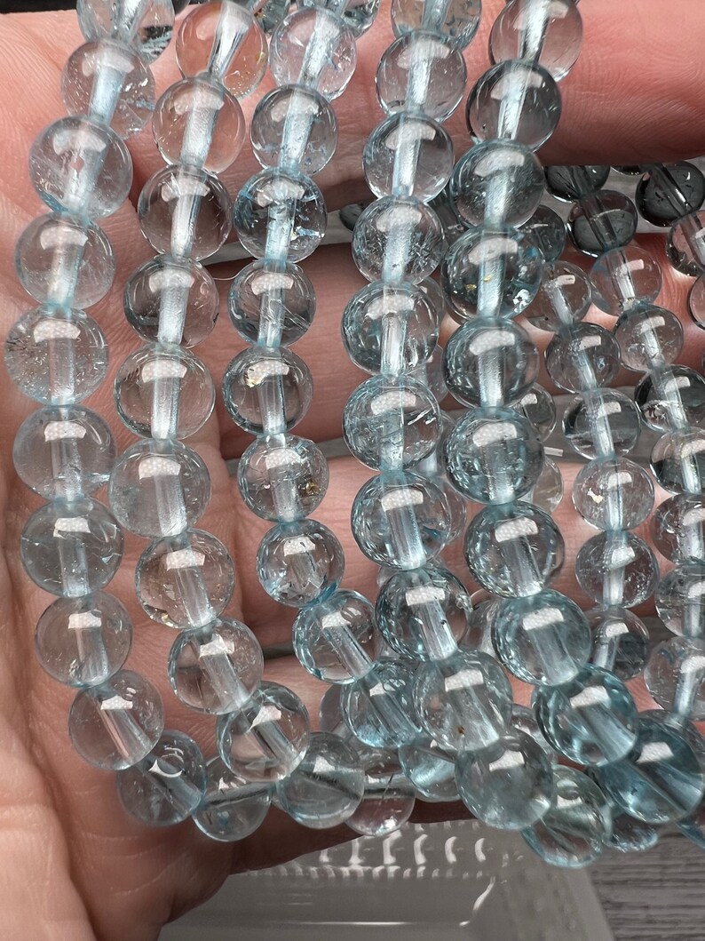 May include: A strand of light blue aquamarine beads, each bead is round and smooth. The beads are strung together on a clear thread.