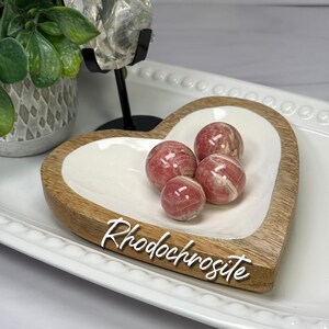 May include: Four polished, spherical rhodochrosite stones with pink and white banded patterns resting inside a heart-shaped dish. The dish features a natural light brown wood rim and a smooth white interior basin with the text Rhodochrosite.