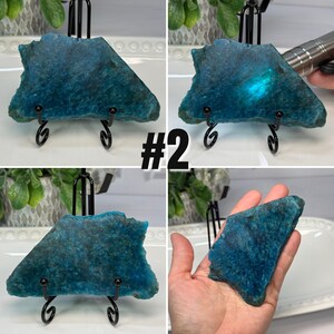 May include: A raw, flat, triangular-shaped blue apatite mineral specimen with a textured surface. The stone displays deep blue and teal hues, with a bright, glowing light reflection visible when illuminated. It is labeled with the text #2.