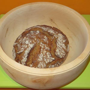 Könnte beinhalten: Eine runde Holzschüssel mit hellbrauner Oberfläche, die ein Brot aus dunklem Braun mit einer flockigen, strukturierten Kruste enthält. Das Brot ist mit kleinen weißen Flocken bedeckt, möglicherweise Haferflocken.