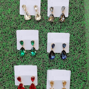 May include: Six pairs of dangle earrings with teardrop-shaped crystals. The earrings are displayed on white cards against a green background. The crystals are in various colors, including clear, gray, green, blue, red, and yellow.