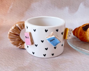 Puede incluir: Taza de cerámica blanca con asa marrón y patrón de corazones negros. La taza tiene tres pequeñas decoraciones en forma de libro con la palabra "Book" en ellas.