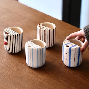 Puede incluir: Cuatro tazas de cerámica con patrones de rayas. Las tazas son blancas con rayas negras, marrones, azules y rojas. Las tazas están sobre una superficie de madera.