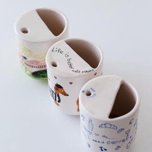 May include: Three white ceramic mugs with different hand-painted designs. The first mug has a colorful landscape scene, the second mug has a dachshund with the text "Life is better with paws", and the third mug has a blue and white design with the text "I need coffee".