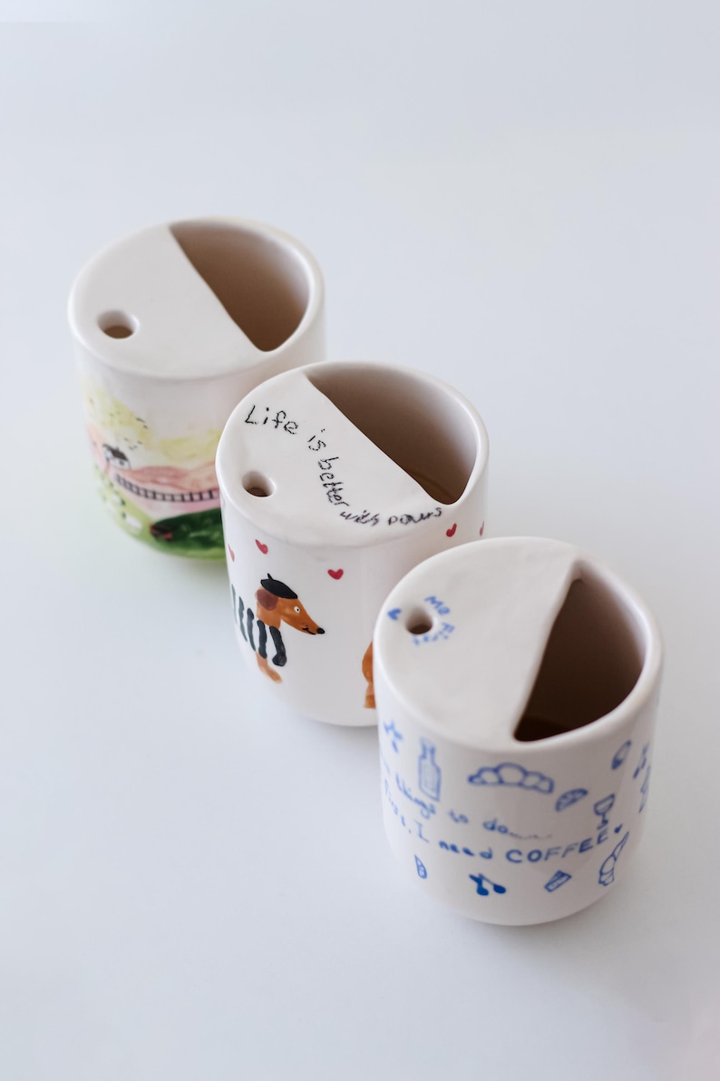 May include: Three white ceramic mugs with different hand-painted designs. The first mug has a colorful landscape scene, the second mug has a dachshund with the text "Life is better with paws", and the third mug has a blue and white design with the text "I need coffee".