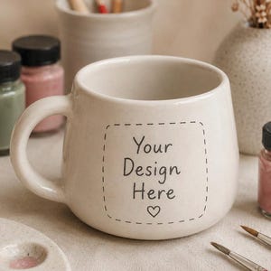 Può includere: Una tazza in ceramica color crema con una forma arrotondata e un singolo manico. La parte anteriore della tazza presenta una cornice quadrata con linee tratteggiate contenente il testo nero "Your Design Here" e un piccolo simbolo a cuore.