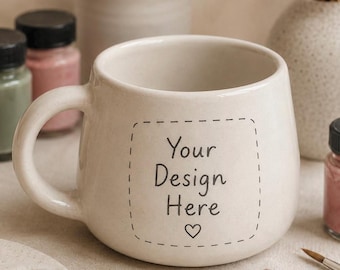 Könnte beinhalten: Ein cremefarbener Keramikbecher mit abgerundeter Form und einem einzelnen Henkel. Auf der Vorderseite des Bechers befindet sich ein quadratischer Rahmen mit gestrichelten Linien, der den schwarzen Text "Your Design Here" und ein Herz enthält.