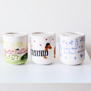May include: Three white ceramic mugs with hand-painted designs. The first mug features a countryside scene with a house, sheep, and a fence. The second mug has a black and white striped dachshund with a beret. The third mug has blue text that reads "I have things to do... But first, I need coffee."