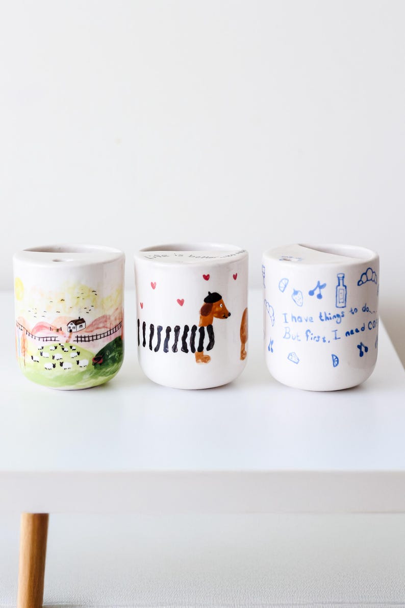May include: Three white ceramic mugs with hand-painted designs. The first mug features a countryside scene with a house, sheep, and a fence. The second mug has a black and white striped dachshund with a beret. The third mug has blue text that reads "I have things to do... But first, I need coffee."
