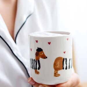 May include: White ceramic mug with a hand-painted design of two dachshunds wearing black and white striped outfits. One dachshund is wearing a beret. The mug has red hearts painted around the rim.