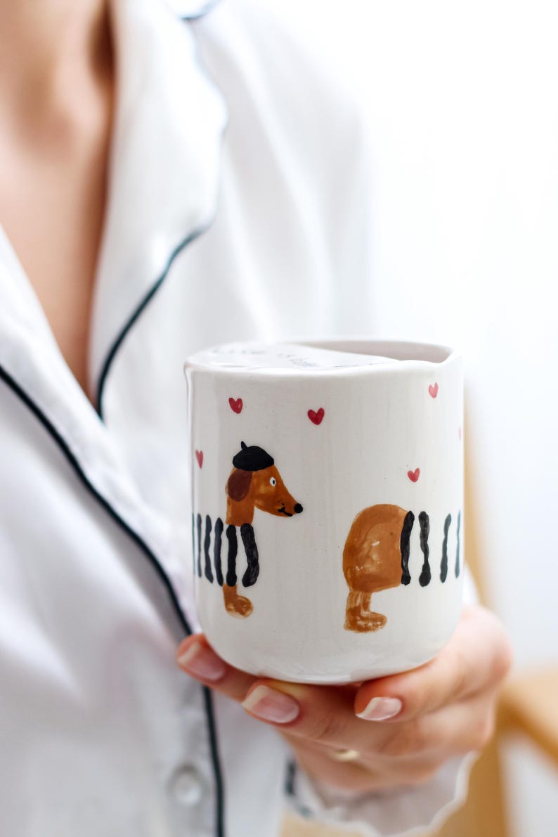 May include: White ceramic mug with a hand-painted design of two dachshunds wearing black and white striped outfits. One dachshund is wearing a beret. The mug has red hearts painted around the rim.