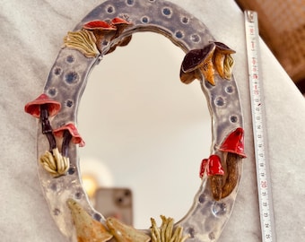 May include: An oval ceramic mirror with a textured, mottled grey frame decorated with three-dimensional sculpted mushrooms in shades of red, brown, and tan. A white measuring tape placed alongside shows the mirror is approximately 12 inches long.