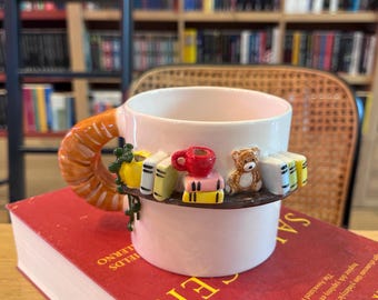 Puede incluir: Una taza de cerámica blanca con un asa decorativa y un diseño de estante. El estante presenta libros en miniatura, una taza roja y un pequeño oso de peluche. La taza está sobre un libro rojo con letras doradas.