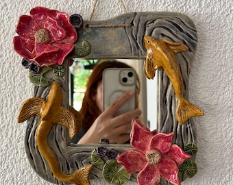 May include: Square ceramic mirror with a gray wood-grain frame, adorned with two golden fish and two red flowers. The mirror hangs from a natural fiber cord and is mounted on a white textured wall.