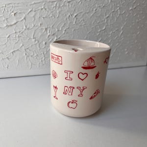Può includere: Una tazza in ceramica color crema con illustrazioni rosse e la scritta "I NY". La tazza presenta disegni di una barca a vela, una mela, una fetta di pizza e altre icone di New York. La tazza è cilindrica e ha una superficie liscia.