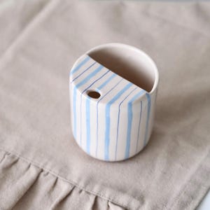 Könnte beinhalten: Kleine, cremefarbene Keramikvase mit runder Form und diagonalem Schnitt auf einer Seite. Die Vase ist mit vertikalen blauen Streifen verziert. Ein kleines Loch ist an der Seite sichtbar. Auf einer beigen Leinenoberfläche.