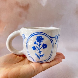 May include: White ceramic mug with a blue floral design. The mug has a textured surface and a rounded handle.