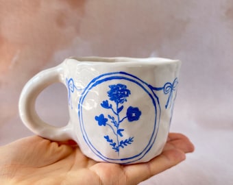 May include: White ceramic mug with a blue floral design. The mug has a textured surface and a rounded handle.