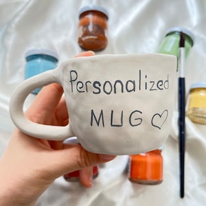 May include: A white ceramic mug with the words "Personalized MUG" and a heart drawn on it. The mug is being held by a hand and there are several small jars of paint in the background.
