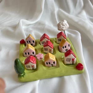 May include: A ceramic game board with a green field and nine small houses with pink and red roofs. The houses have white windows and a small heart on the front. There is a small tree and a cloud on the board.
