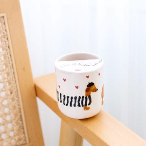 May include: White ceramic mug with a black and white striped dachshund wearing a beret and a red heart. The mug has the text "Life is better with dogs" and red hearts around the dachshund.