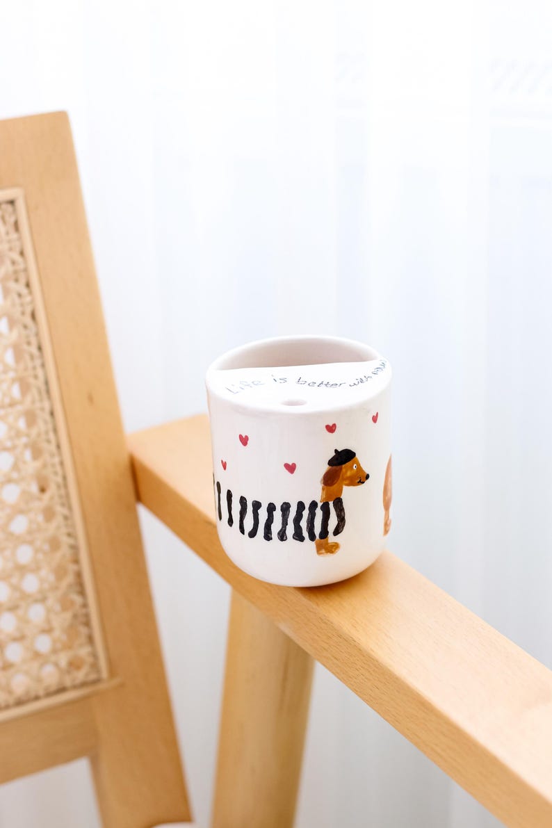 May include: White ceramic mug with a black and white striped dachshund wearing a beret and a red heart. The mug has the text "Life is better with dogs" and red hearts around the dachshund.