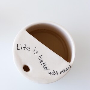 May include: White ceramic mug with a divided section and a hole in the bottom. The mug has the handwritten message "Life is better with paws" on the side.
