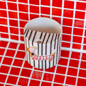 May include: A white ceramic wall pocket with black and white stripes and a dachshund design. The dachshund wears a pink and red striped outfit and holds a green ice cream cone. The pocket is mounted on a red tile background.