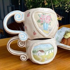 Pode incluir: Duas canecas de cerâmica com designs caprichosos. A caneca superior apresenta um design em forma de coração com um buquê de rosas cor-de-rosa em um vaso azul. A caneca inferior tem uma cena emoldurada de uma casa branca com um gramado verde e um céu azul.