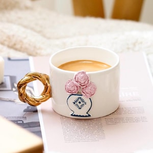 May include: White ceramic coffee mug with a gold-colored twisted handle. The mug features a decorative vase design with pink roses. The mug is filled with coffee and rests on a magazine.